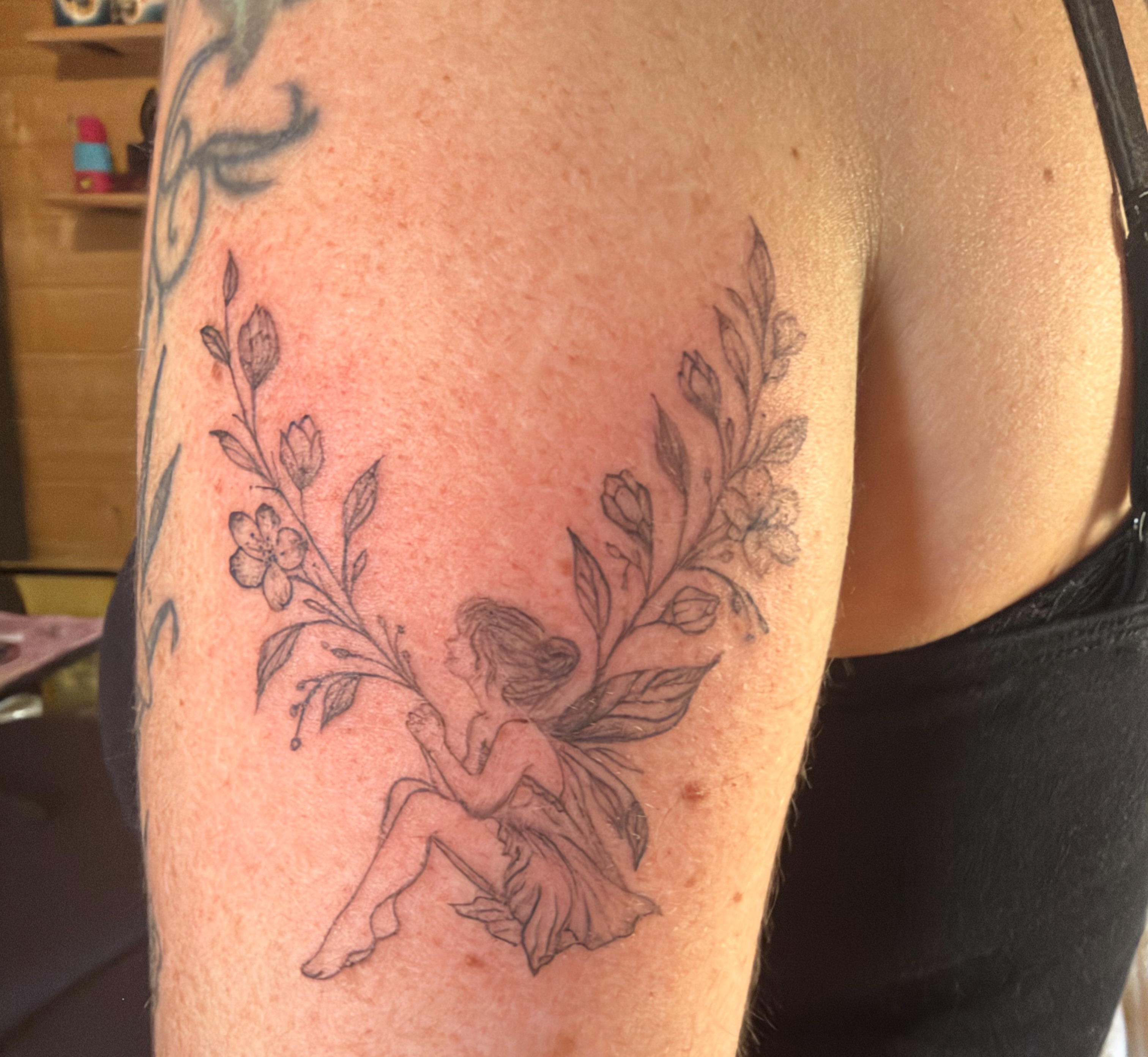 Fine line fairy among floral sprays on an upper arm
